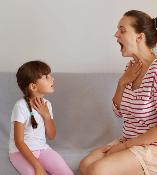 Side view portrait of speech pathologist demonstrating for little kid how to pronounce sounds right, Professional physiotherapist working on speech defects with small child girl indoors.